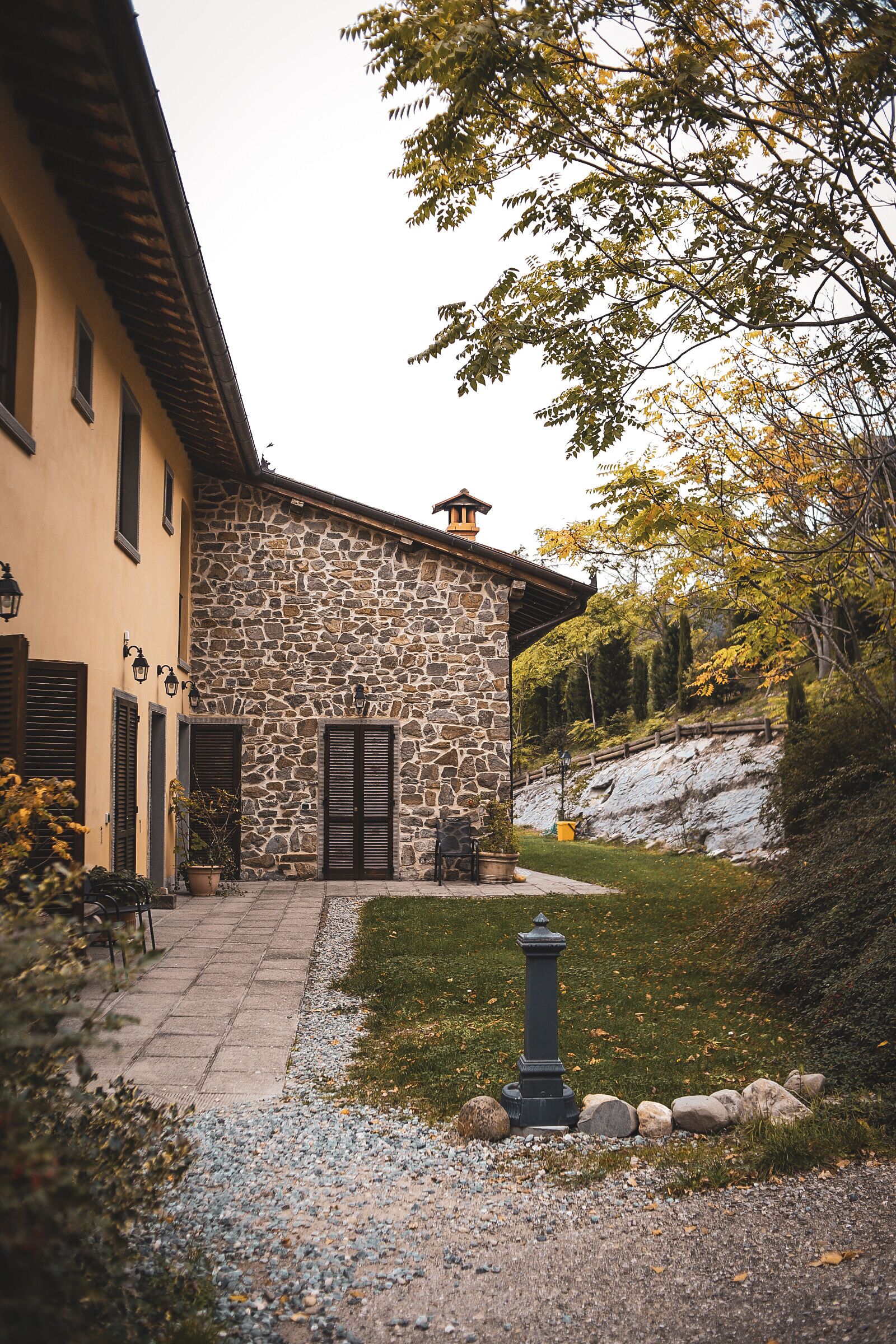 Un edificio de piedra con fachada de piedra y puerta de madera. Un camino empedrado conduce a la entrada, y un pequeño jardín con una farola y rocas.
