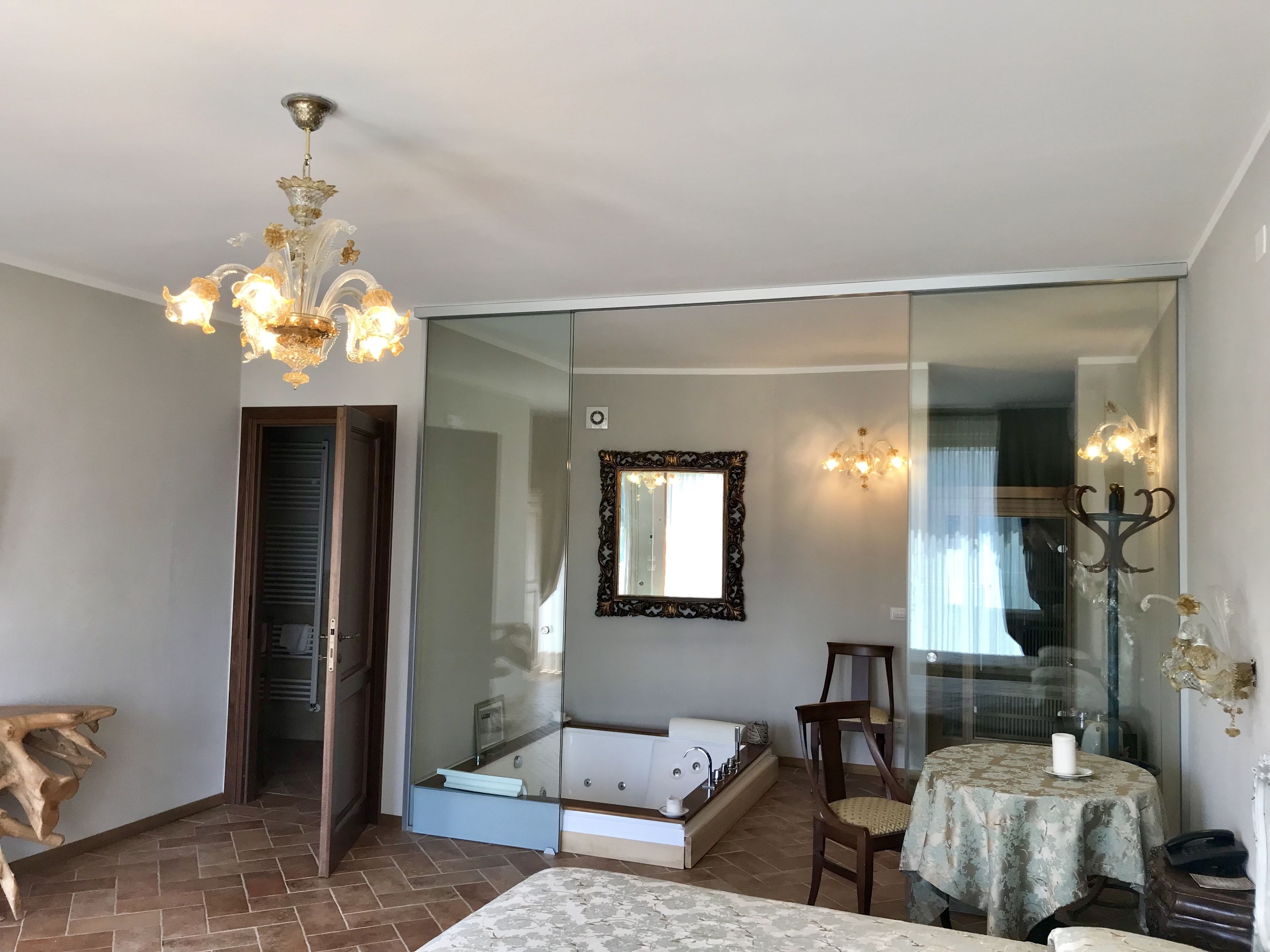 A hotel room with a large mirror, a chandelier, a bathtub, a dining table, and a wooden chair.