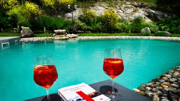 A poolside setting with two glasses of red wine, a book, and a menu on a table. - Borgo I Tre Baroni (Poppi)