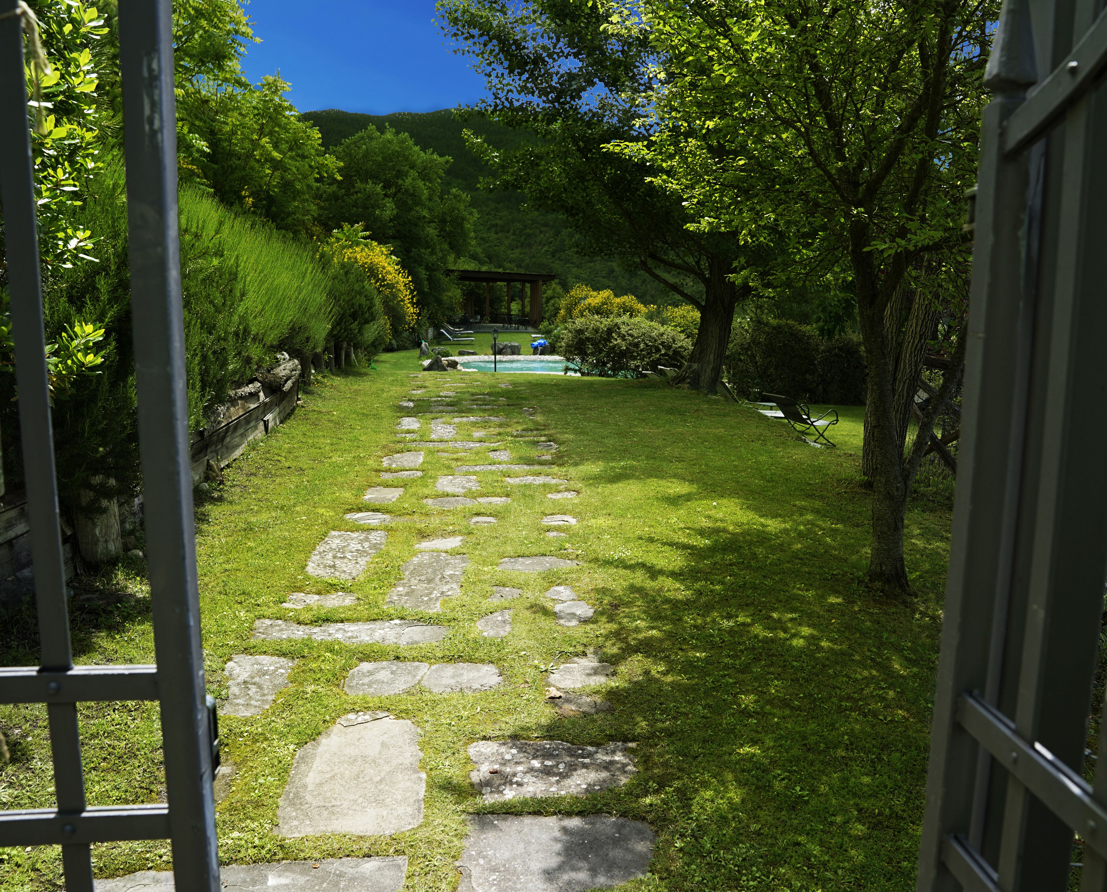 Un sendero de piedra que atraviesa un exuberante jardín con un pabellón y una piscina al fondo.