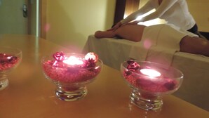 Quarto com uma mesa exibindo duas tigelas de vidro contendo objetos cor-de-rosa, uma cama com uma pessoa sentada e um espelho refletindo a cena.