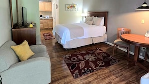 A compact hotel room with a bed, a small sofa, a dining table, and a kitchen area in the background. - Affordable Suites of America Harrisonburg (Harrisonburg)