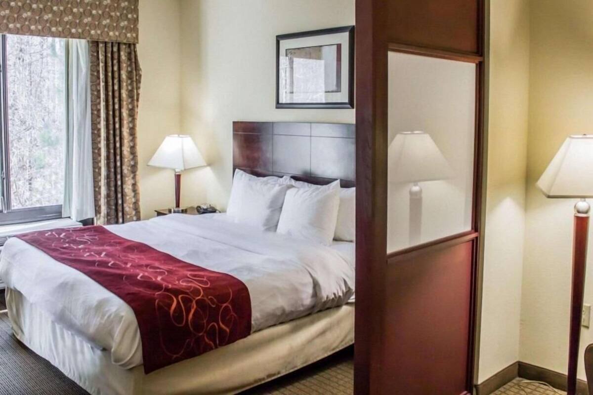 A hotel room with a bed, two bedside lamps, a window with curtains, and a framed picture on the wall.