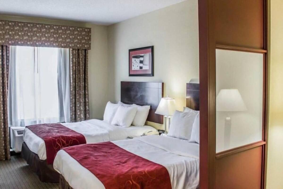A hotel room with two beds, a headboard, a bedside table with a lamp, a window with curtains, and a framed picture on the wall.