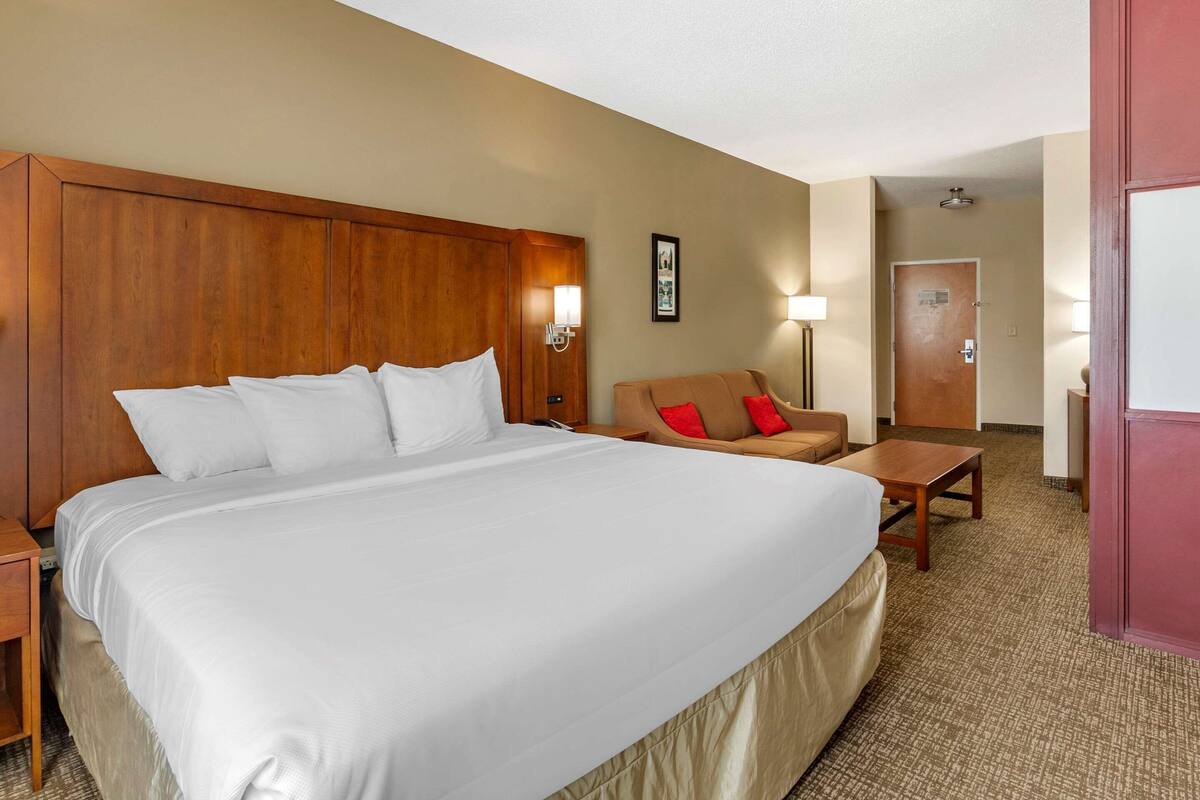 A hotel room with a large bed, a wooden headboard, a sofa with red cushions, a small table, and a lamp.