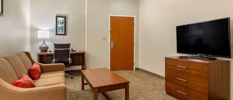 Une chambre d’hôtel avec un canapé, une table en bois, un téléviseur posé sur une commode, un bureau avec une lampe et une porte.