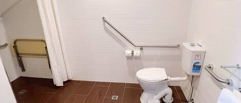 Un baño pequeño y limpio con inodoro blanco, zona de ducha con cortina blanca y barra de apoyo cerca del inodoro.