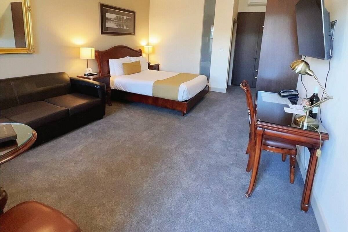 A hotel room with a bed, a sofa, a dining table with chairs, a TV, and a mirror.
