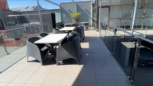 An outdoor terrace with a covered seating area, including tables and chairs, and a glass railing.