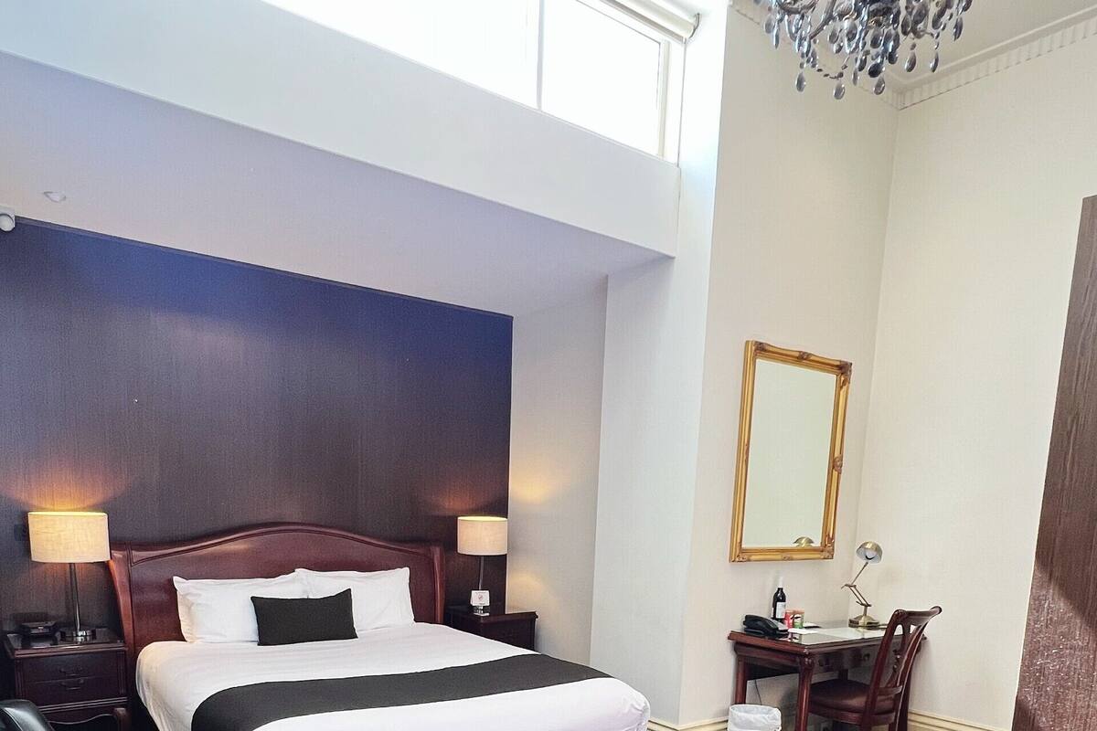 A hotel room with a large bed, a desk, a chair, a mirror, and a chandelier.