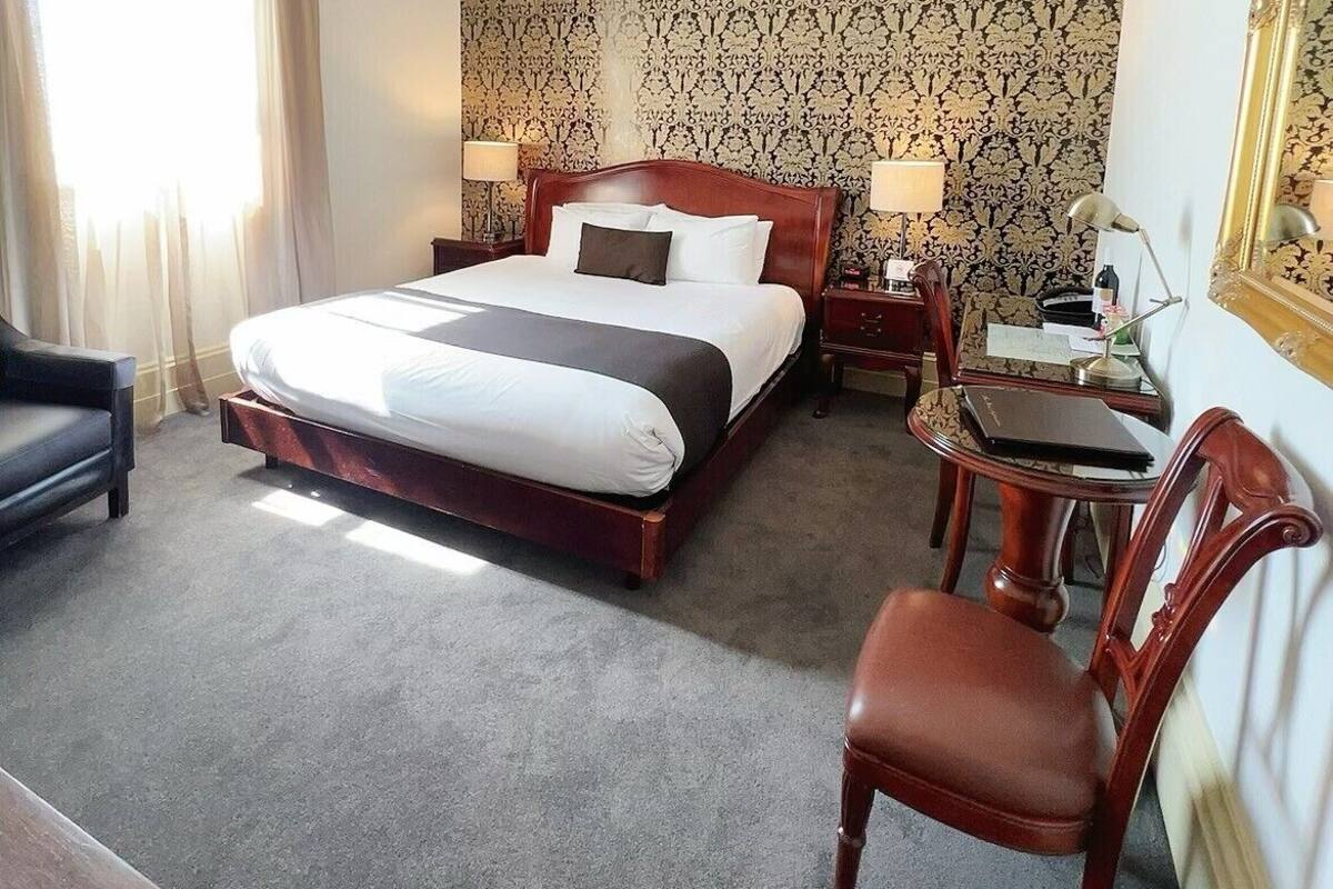 A hotel room with a large bed, a desk with a chair, a chandelier, and a patterned wallpaper.