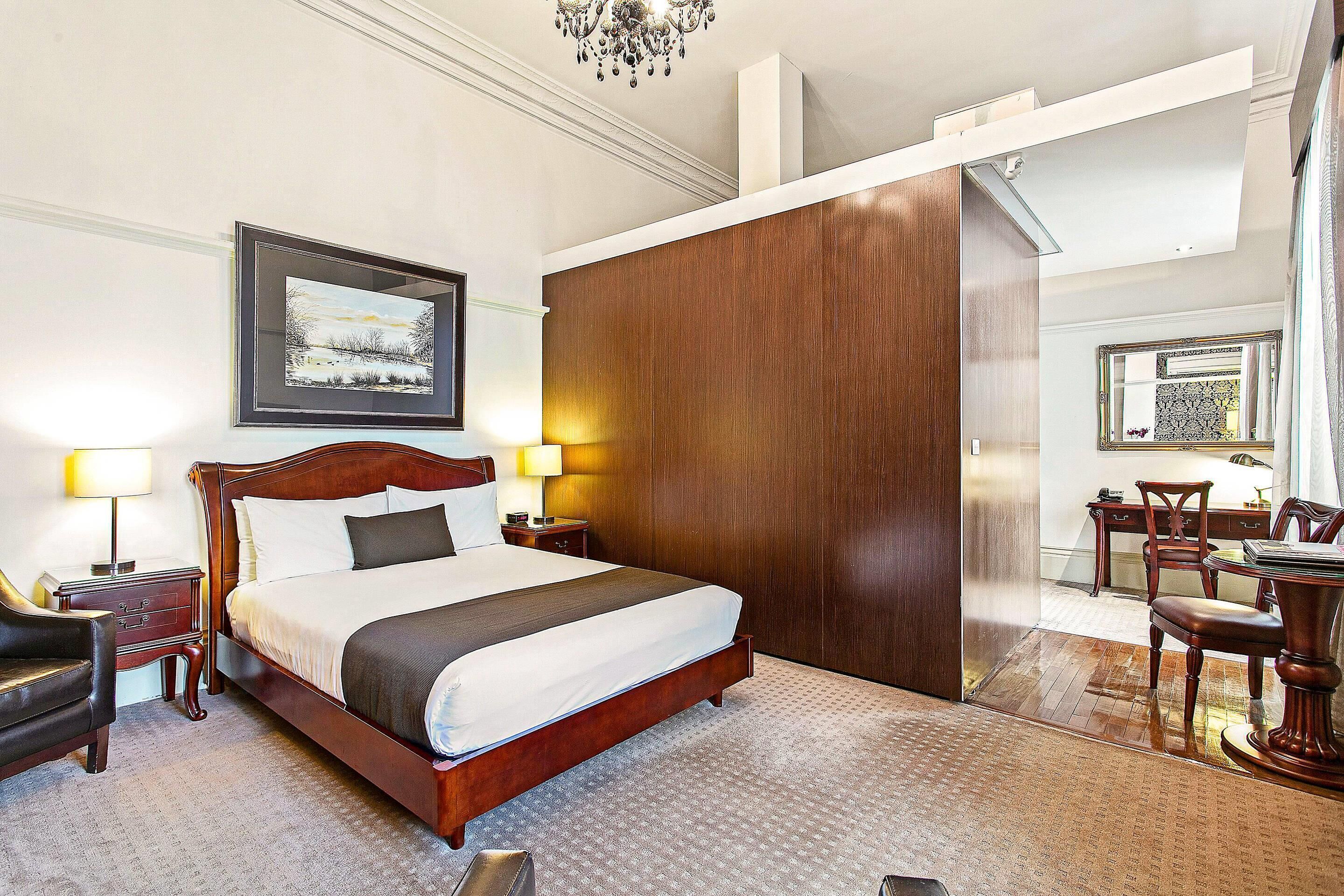 Quarto de hotel com uma cama grande, um guarda-roupa de madeira, uma escrivaninha com cadeira e um candelabro.