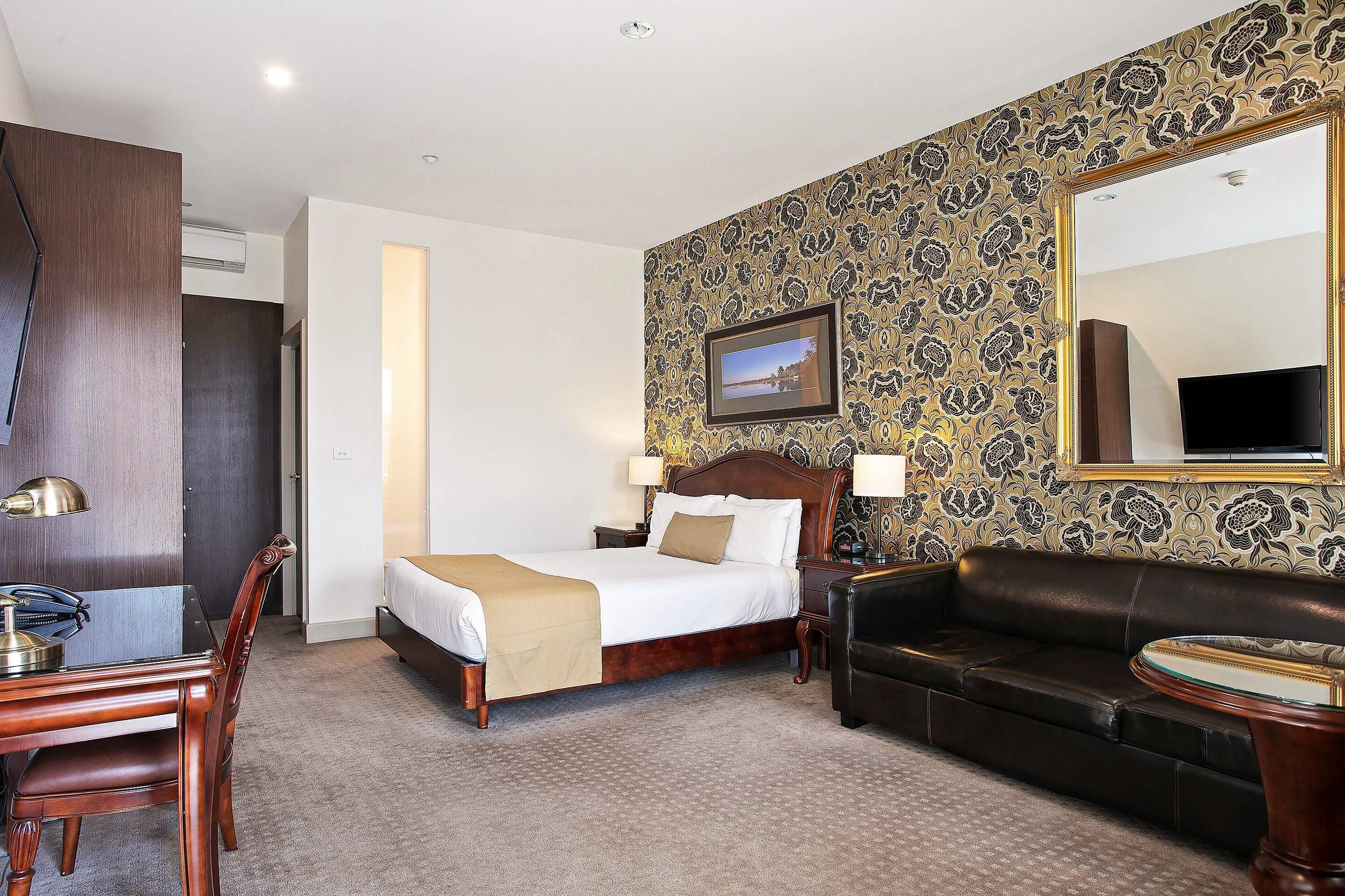 Une chambre d’hôtel comprenant un lit, un canapé, un bureau avec une chaise, un miroir et un mur orné d’un motif floral.