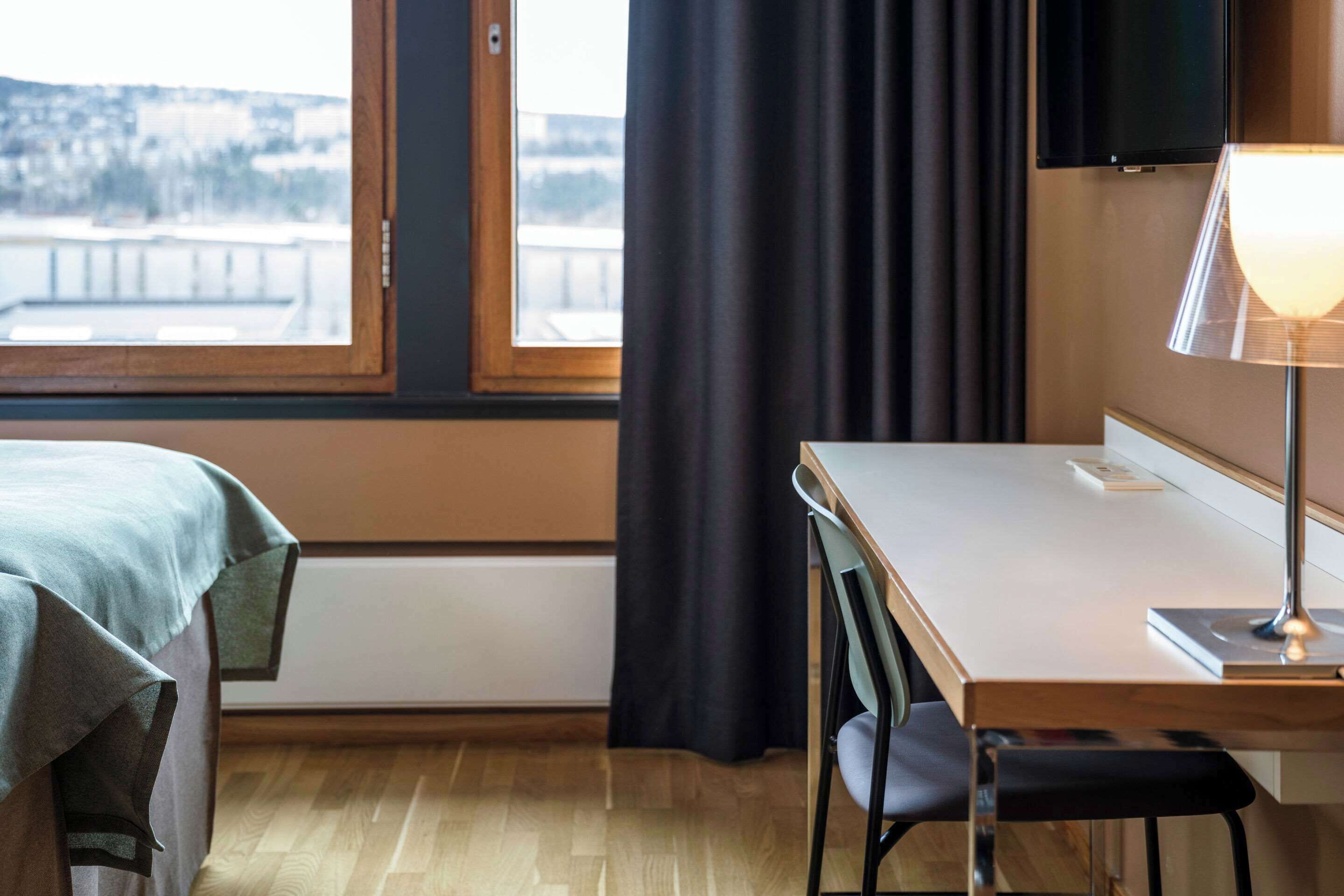 A hotel room with a bed, a desk, a chair, a lamp, a window with curtains, and a view of a bridge.
