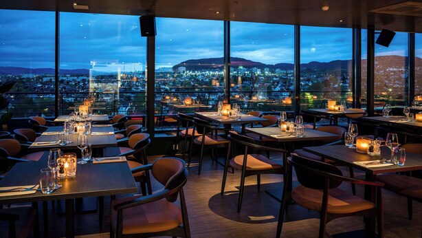 A restaurant with tables set for dining, large windows offering a city view, and candles on the tables. - Quality Hotel 33 (Oslo)