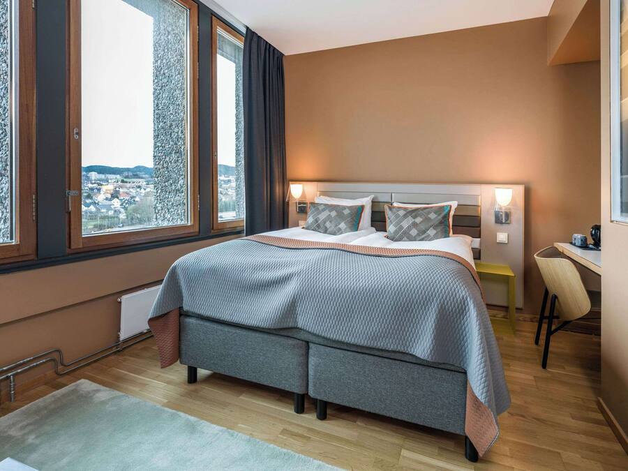 A modern bedroom with a large bed, a desk, and a view of a cityscape through large windows.