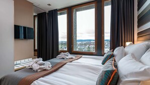 A hotel room with a large bed, a flat-screen TV mounted on the wall, and a view of a cityscape through large windows. - Quality Hotel 33 (Oslo)