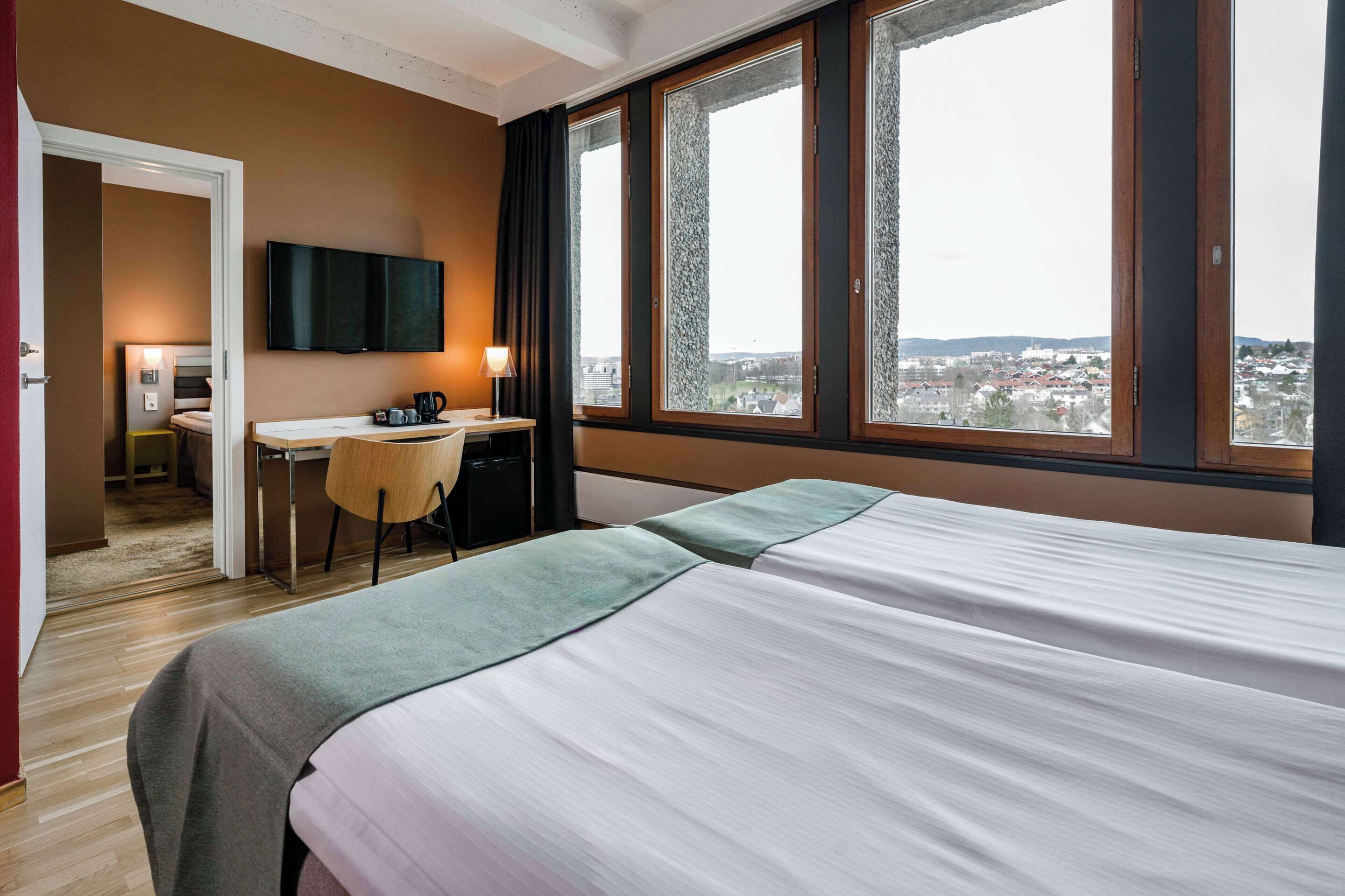 A hotel room with a bed, a desk, a chair, a TV, and large windows offering a city view.