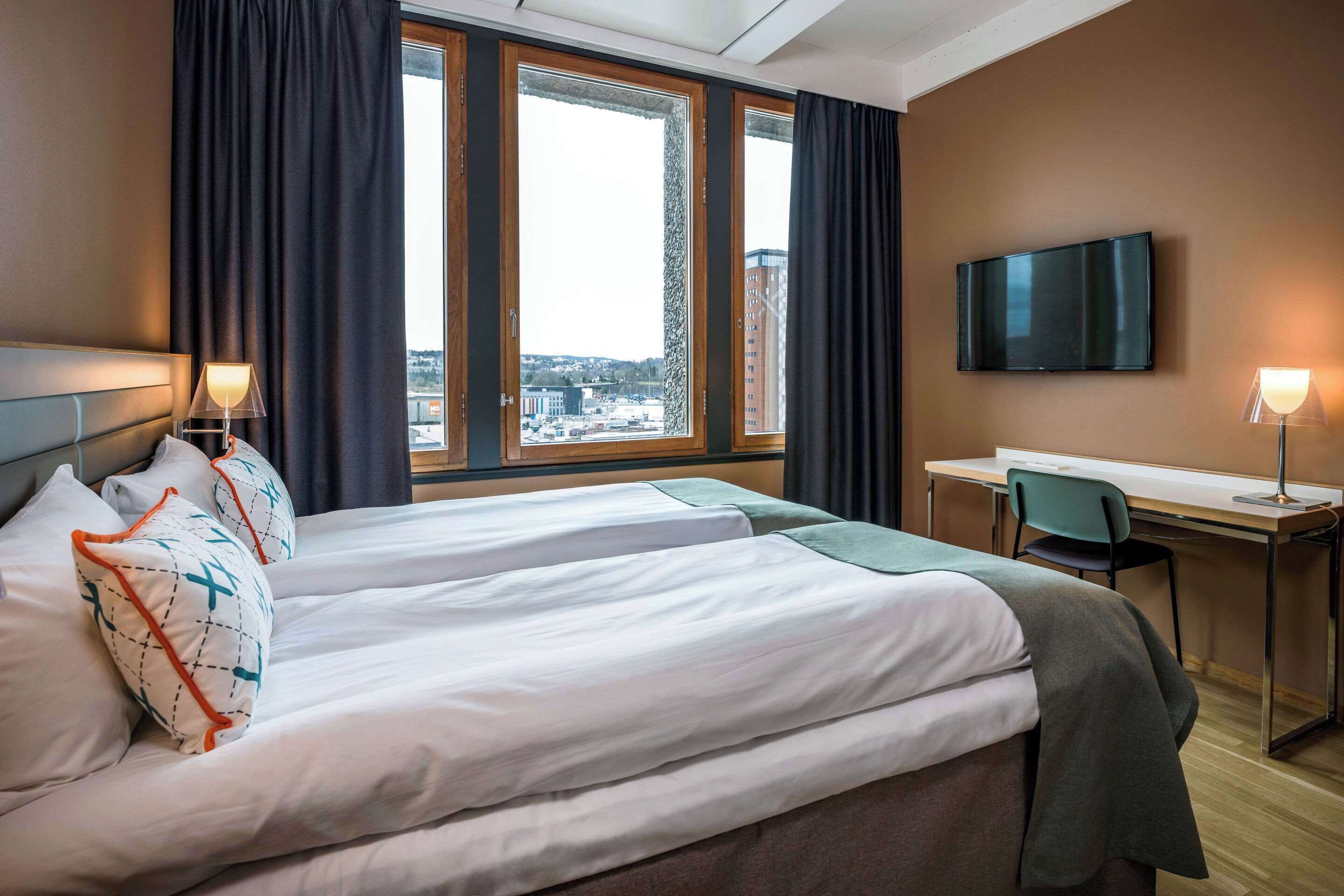 Hotelkamer met twee bedden, een bureau, een stoel, een televisie en uitzicht op de stad door grote ramen.