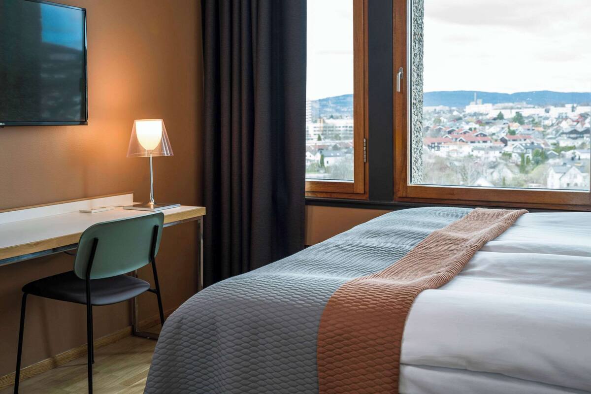 A hotel room with a bed, a desk, a chair, a TV, and a window with a city view.