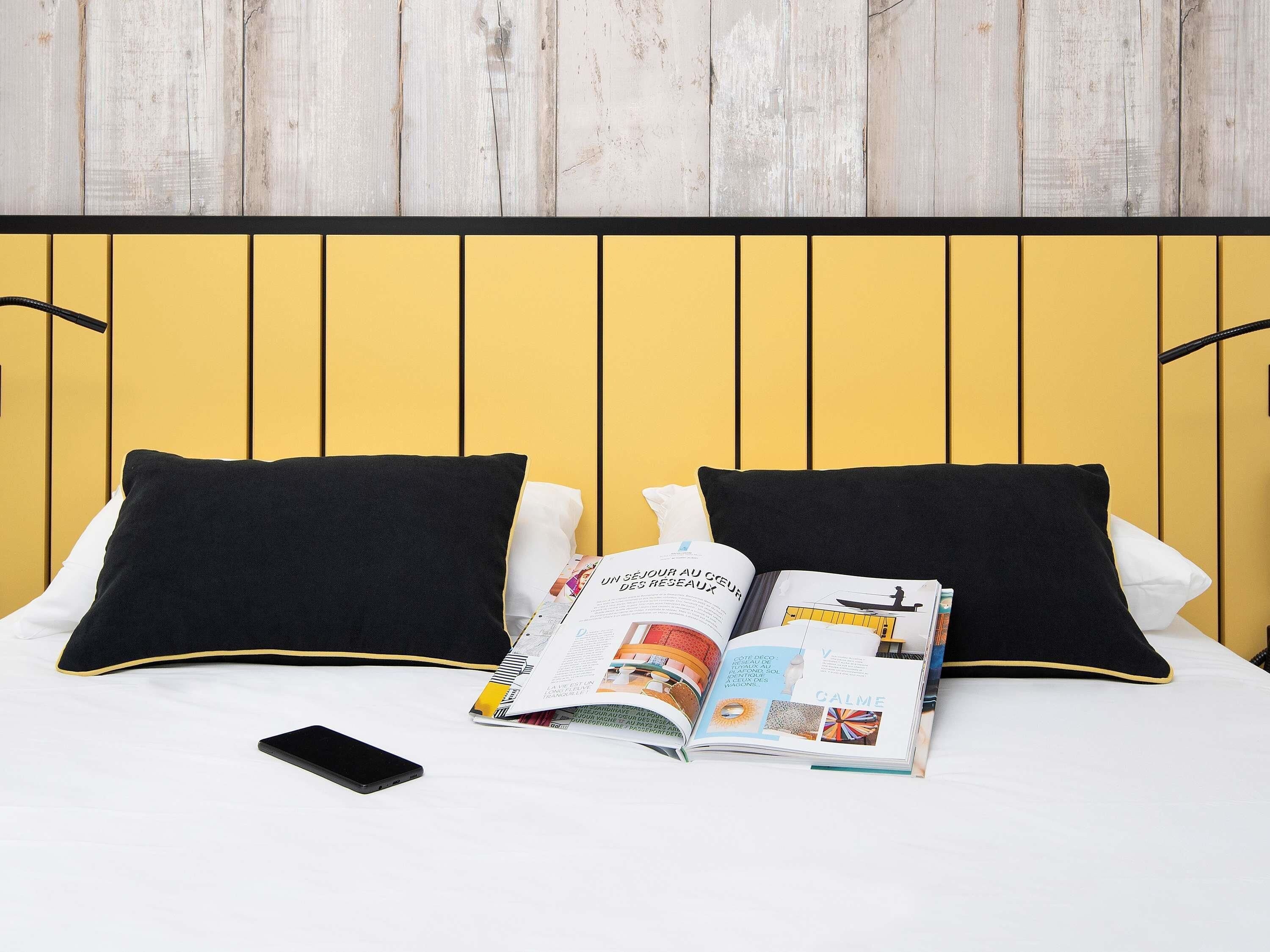 Una cama con almohadas blanco y negro, una revista y un teléfono inteligente sobre un edredón blanco.