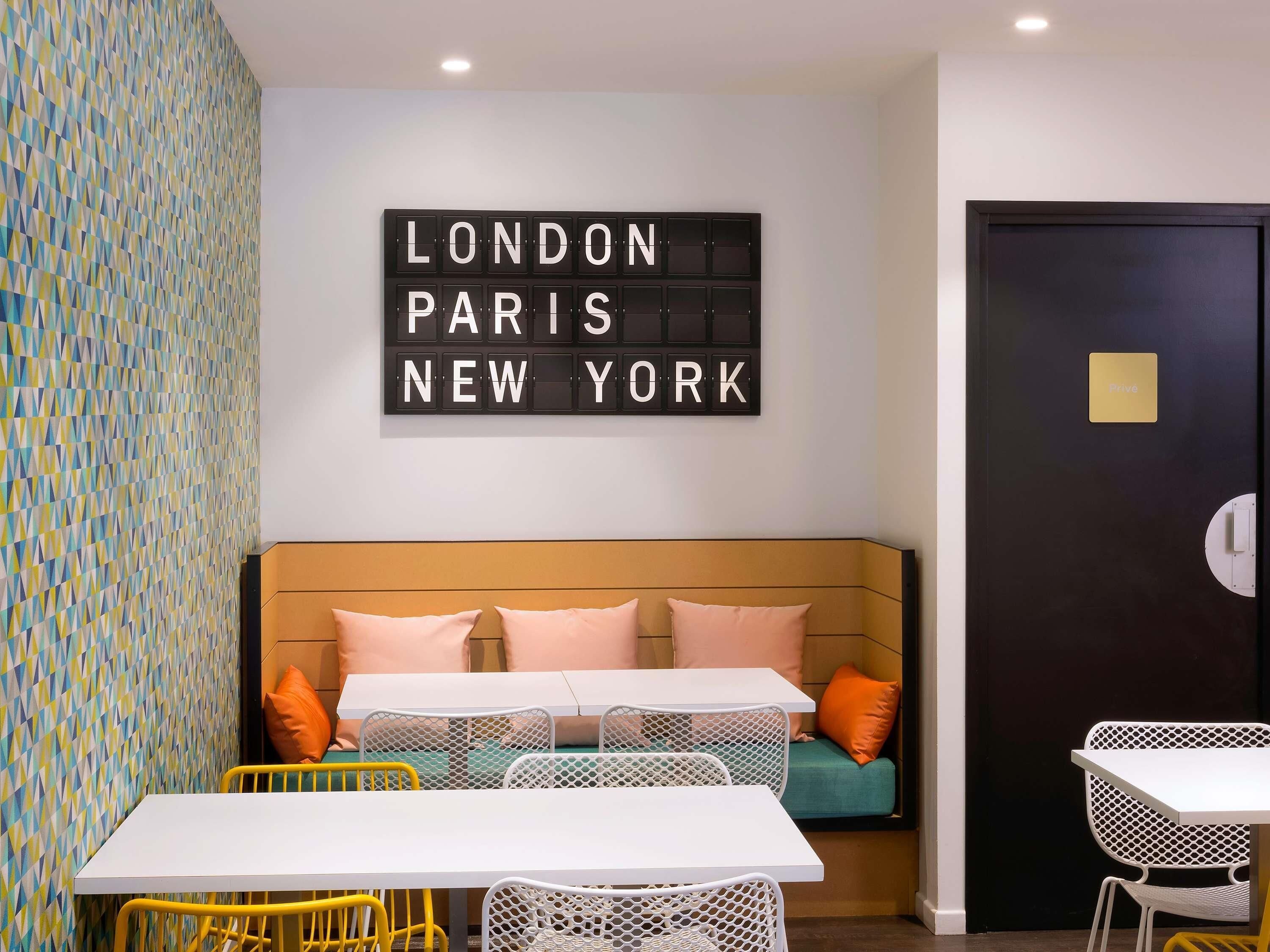 A room with a bed, a table, and chairs, a wall with a geometric pattern, and a sign with city names.