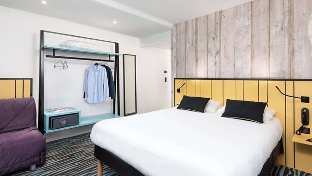 Une chambre d’hôtel moderne avec un grand lit, une armoire intégrée, un coin salon avec un fauteuil violet et un mur d’accent en bois.
