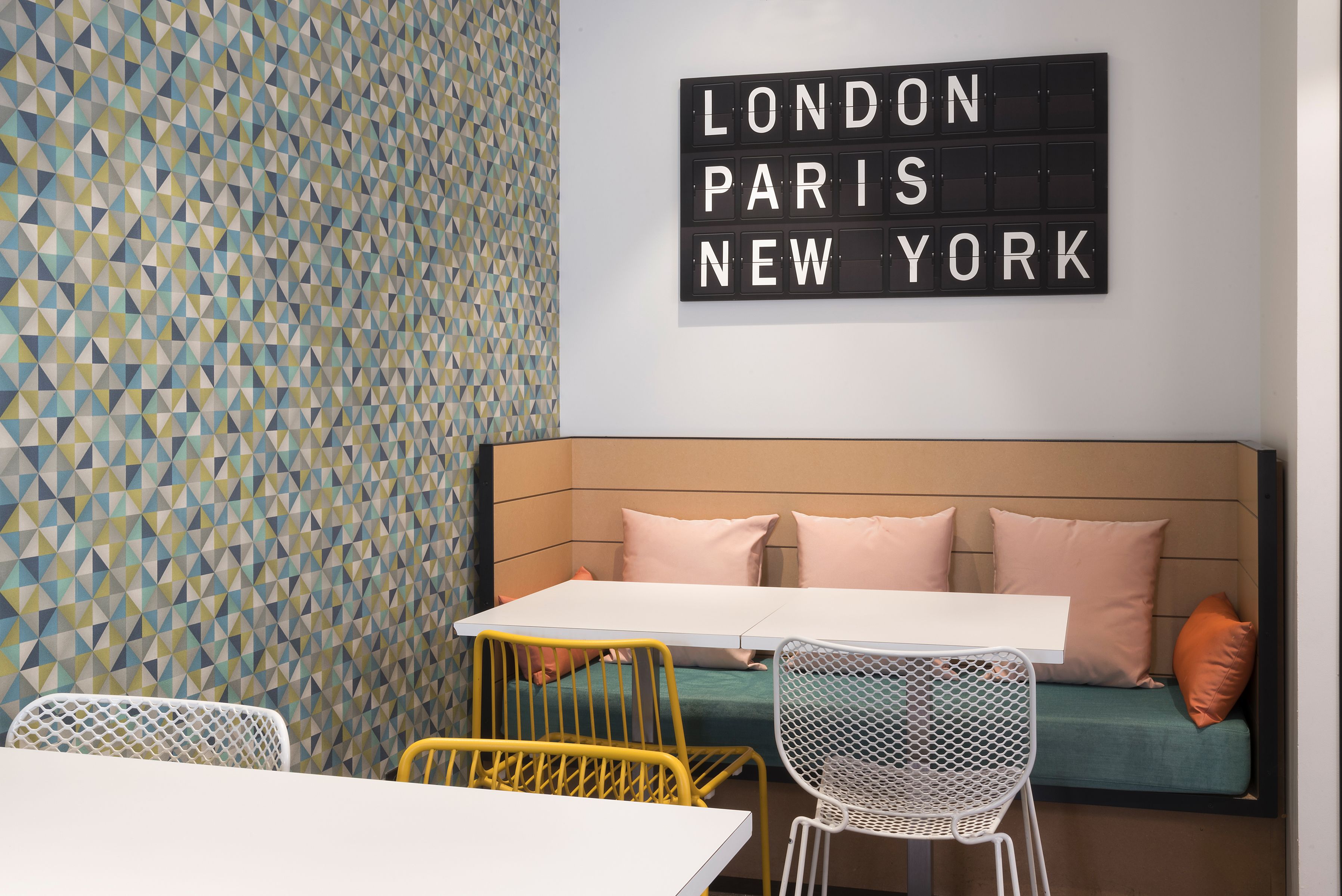 A modern room with a geometric patterned wall, a white table, yellow and white chairs, a green sofa with pink cushions, and a sign reading "LONDON PARIS NEW YORK".