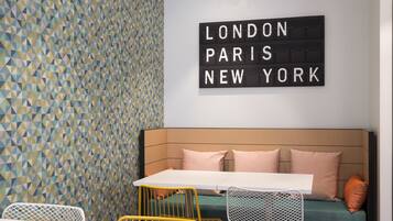 A modern room with a geometric patterned wall, a white table, yellow and white chairs, a green sofa with pink cushions, and a sign reading "LONDON PARIS NEW YORK".