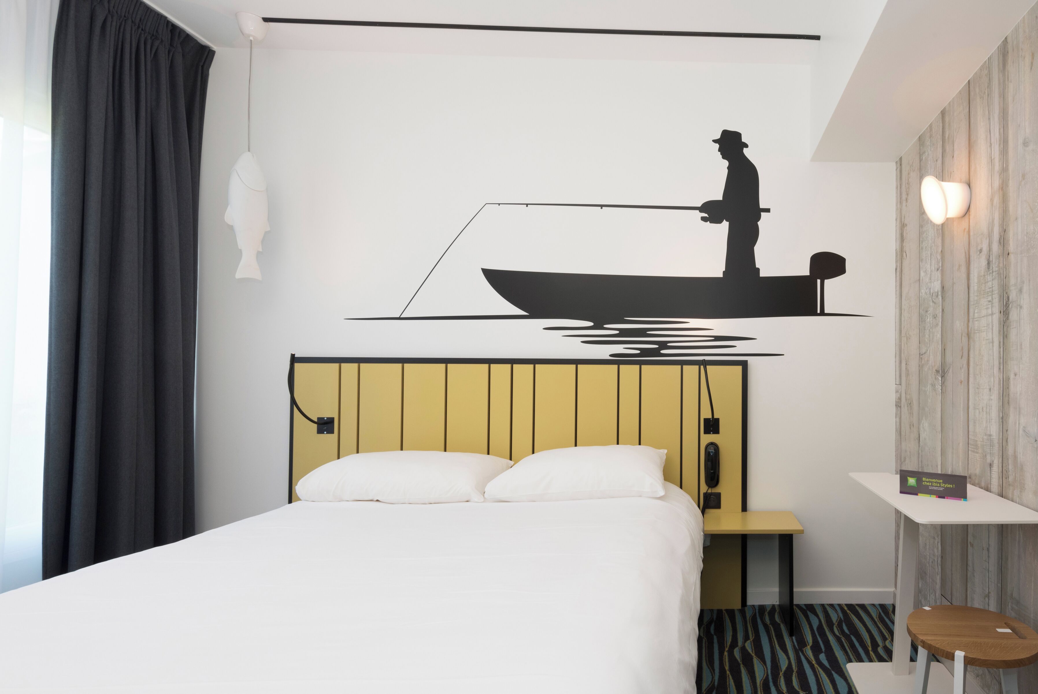 A hotel room with a bed, a nightstand, a wall-mounted lamp, and a decorative wall art of a fisherman.
