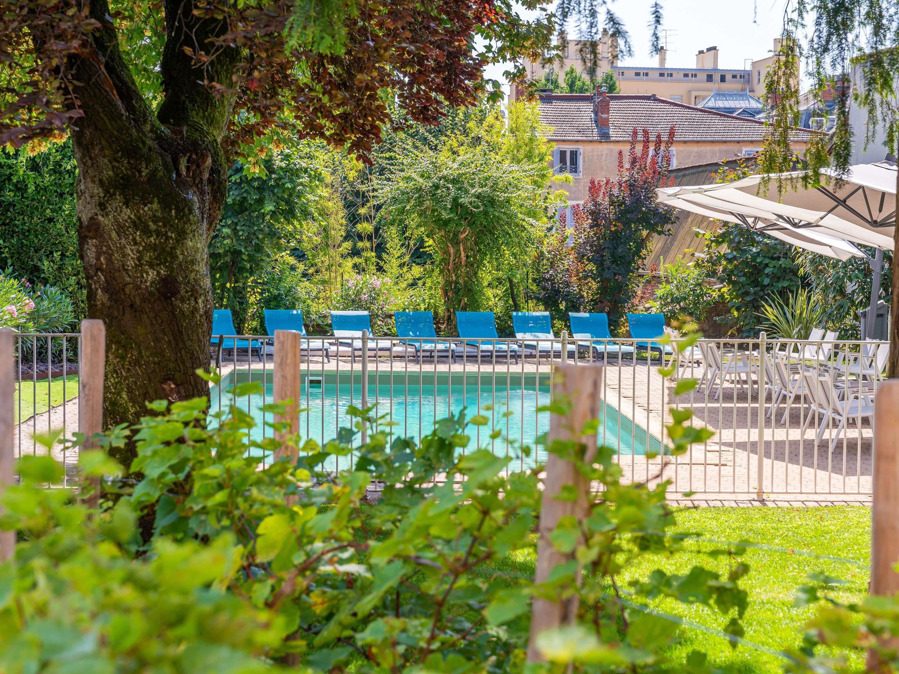 Zona de piscina con sillones azules, un árbol y un edificio al fondo.