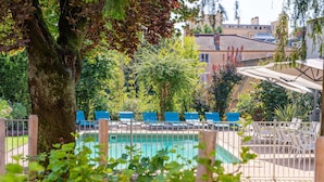 Une zone avec piscine, des chaises longues bleues, un arbre et un bâtiment en arrière-plan.