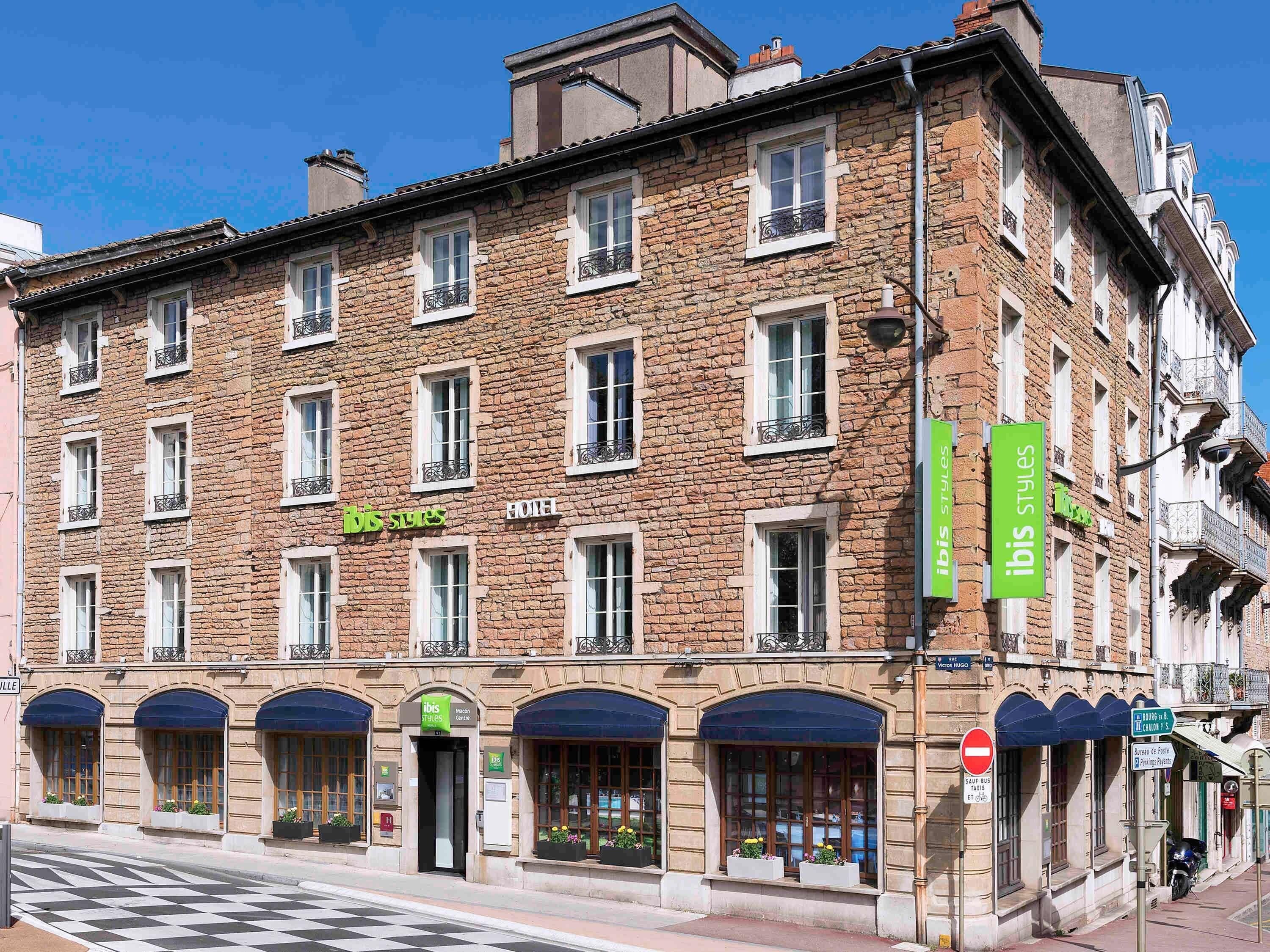 Un hotel de varios pisos con fachada de piedra, múltiples ventanas y un letrero verde 'ibis Styles'.
