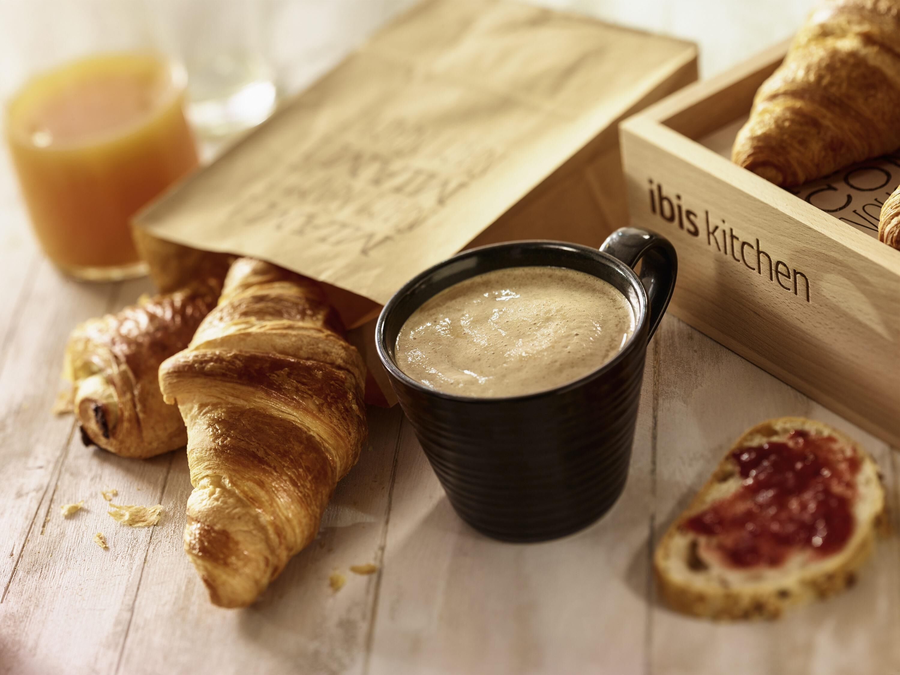 En træbakke med croissanter, en skål suppe og et glas appelsinjuice på en træoverflade.