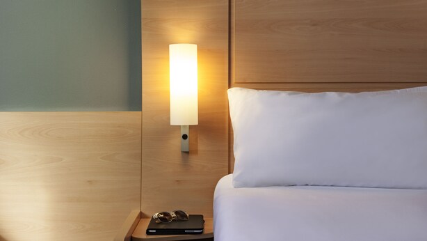 Una cama bien tendida con sábanas blancas y un cabecero de madera. Se ve una mesita de noche con una lámpara y un par de gafas.