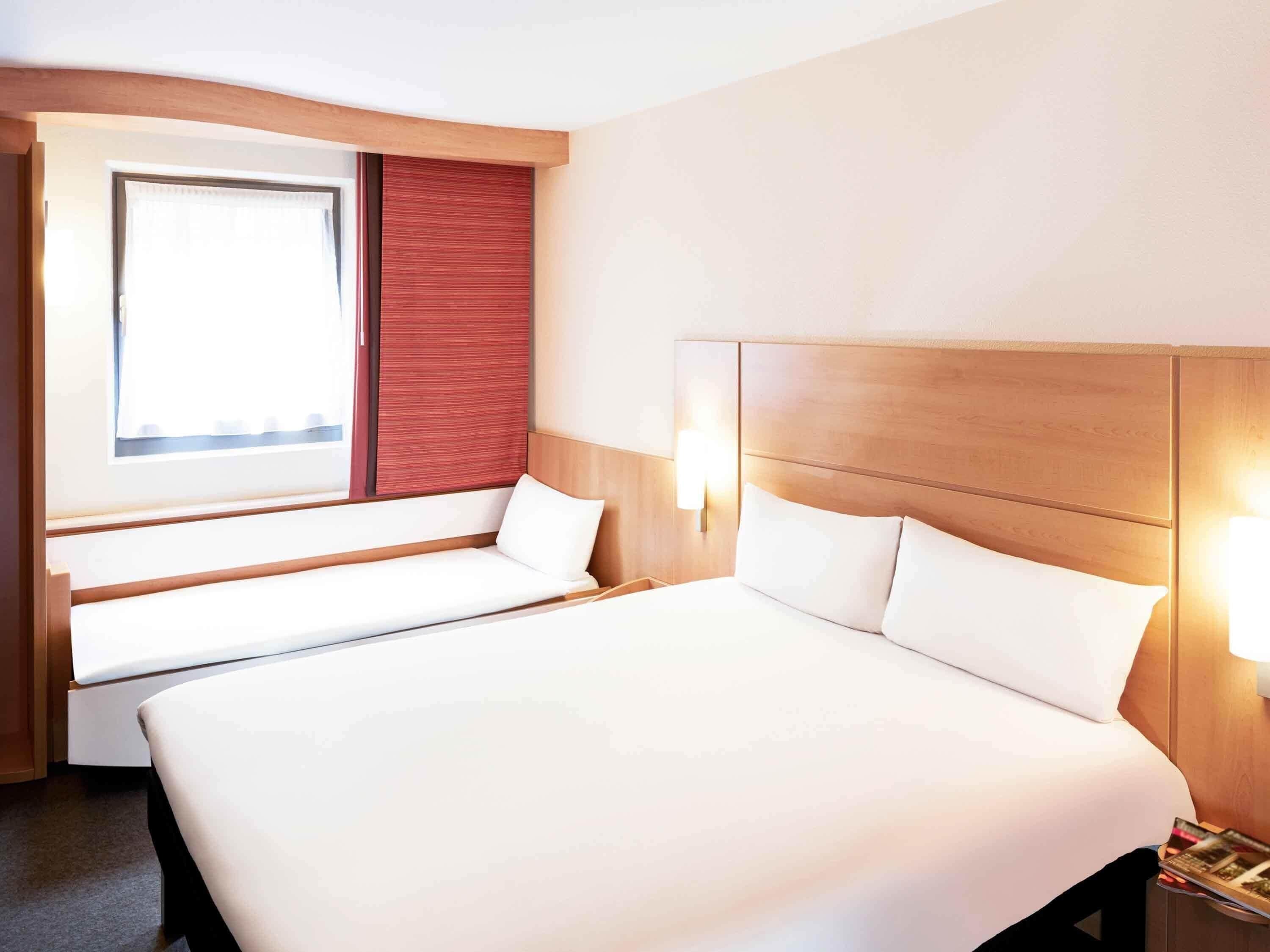 A hotel room with a large bed, a window with a view, a bedside table with a lamp, and a bench by the window.