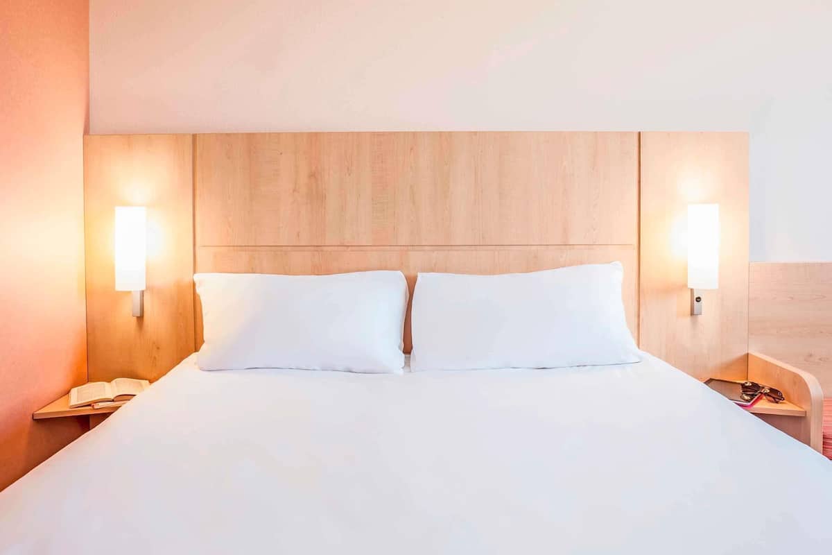 A hotel room with a wooden headboard, two bedside lamps, a white bedspread, and a nightstand with a book and a phone.