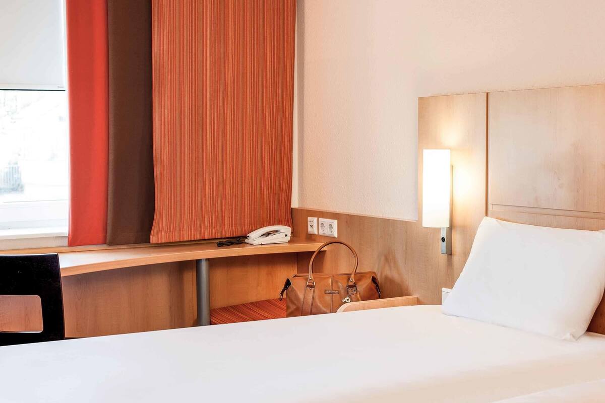 A hotel room with a bed, a desk with a phone, a handbag, and a wall-mounted light.
