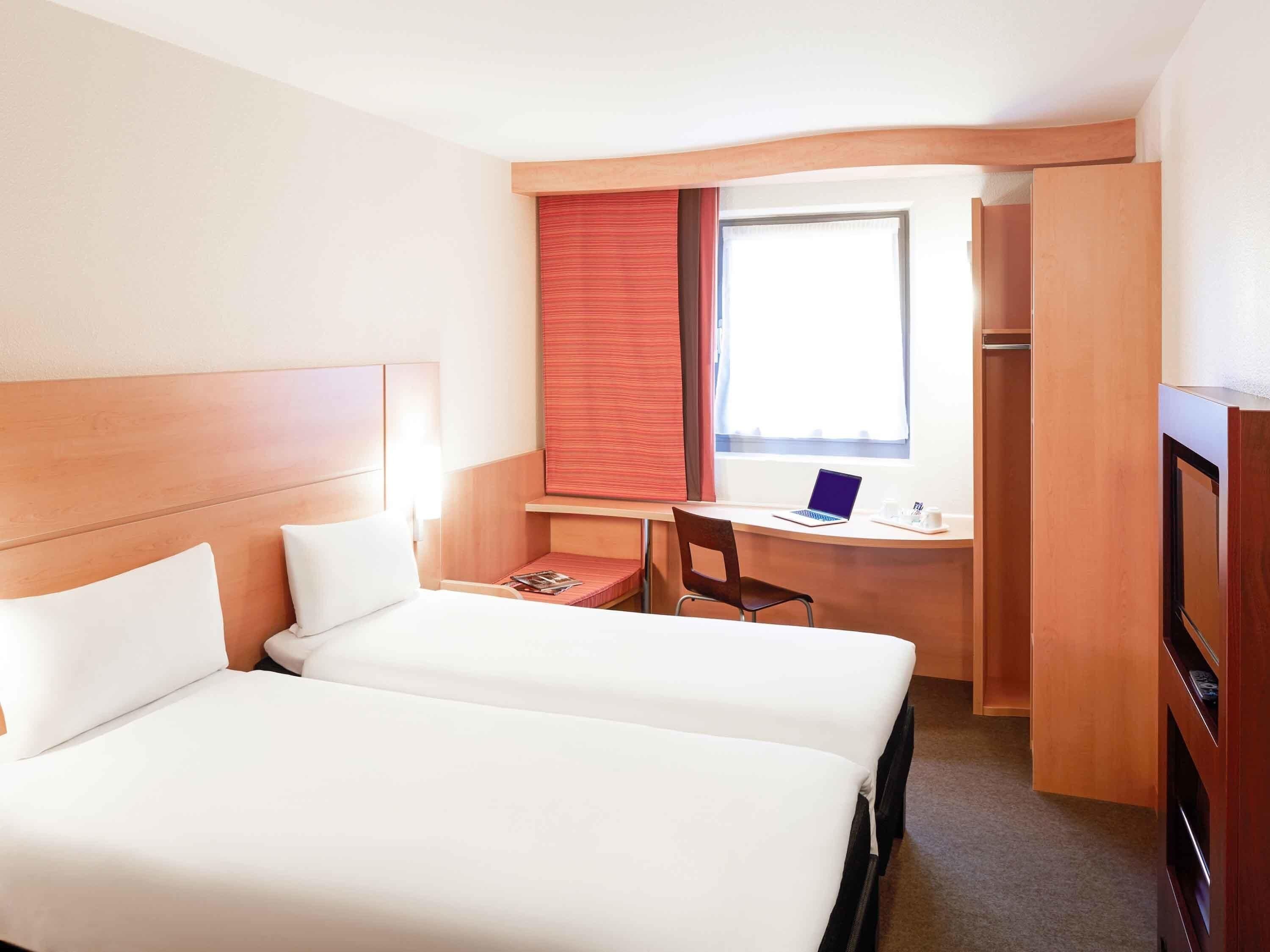 A hotel room with two beds, a desk with a laptop, a chair, and a wardrobe.