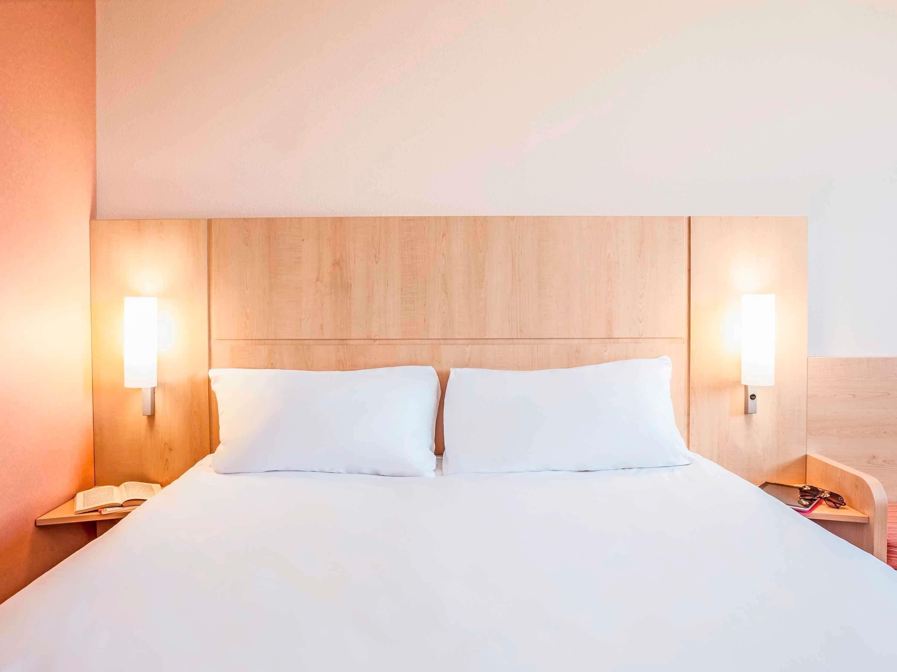 A hotel room with a wooden headboard, two bedside lamps, a white bedspread, and a nightstand with a book and a phone.