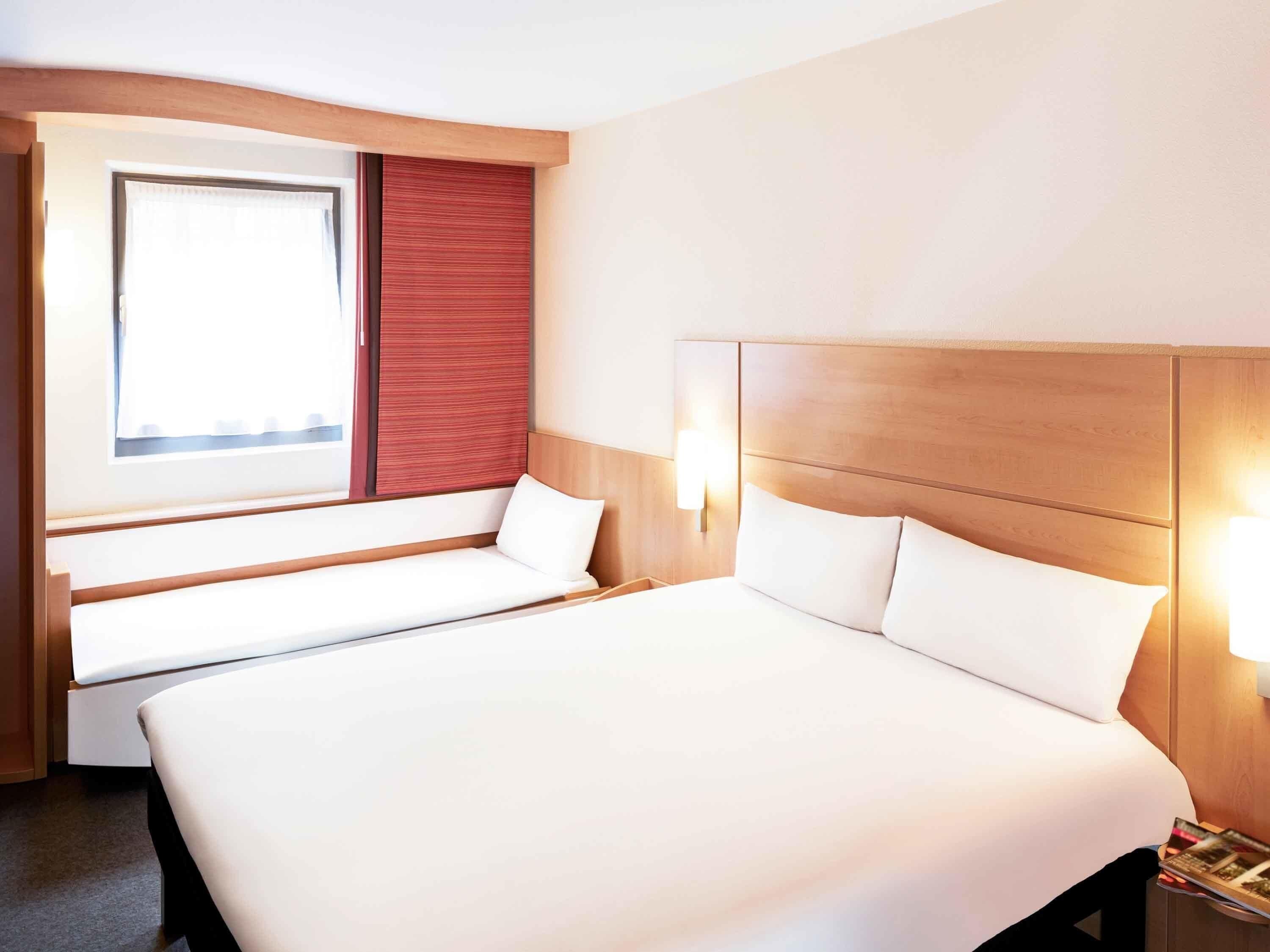 A hotel room with a large bed, a window with a view, a bedside table with a lamp, and a bench by the window.