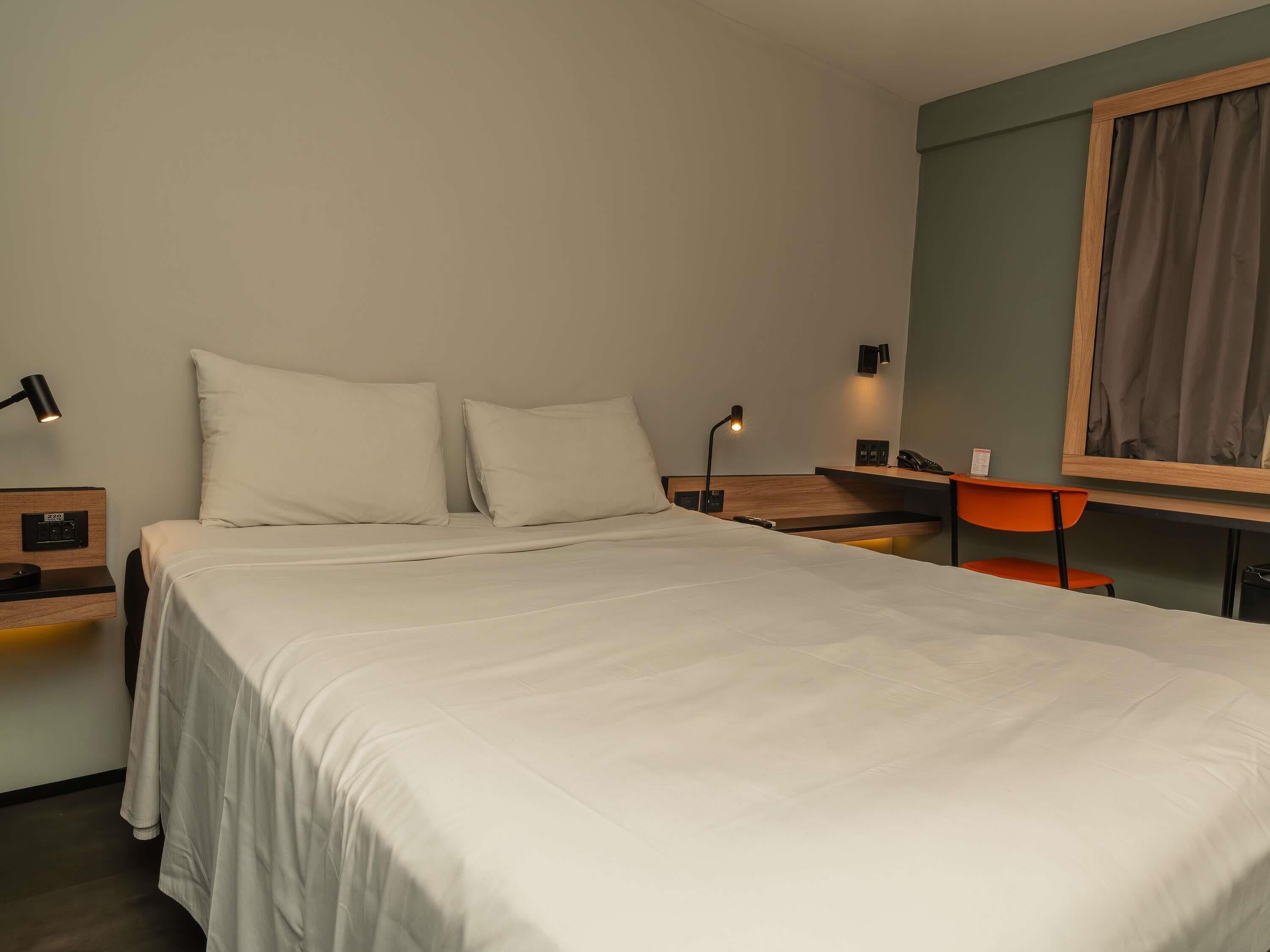 Una cama bien hecha con ropa blanca, una mesita de noche con lámpara, un escritorio pequeño con silla y un espejo montado en la pared.