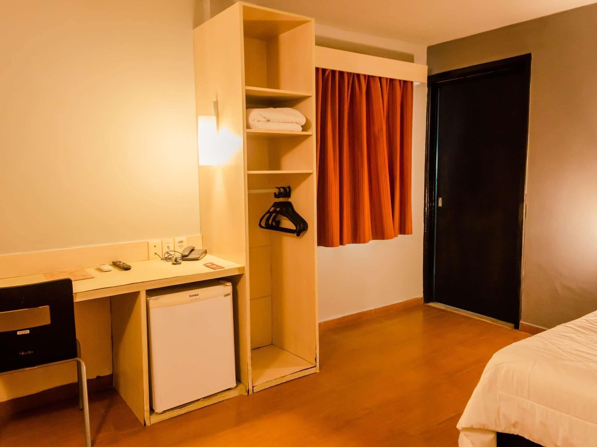 Hotellihuone, jossa on työpöytä, pieni jääkaappi, vaatekaappi ja sänky.