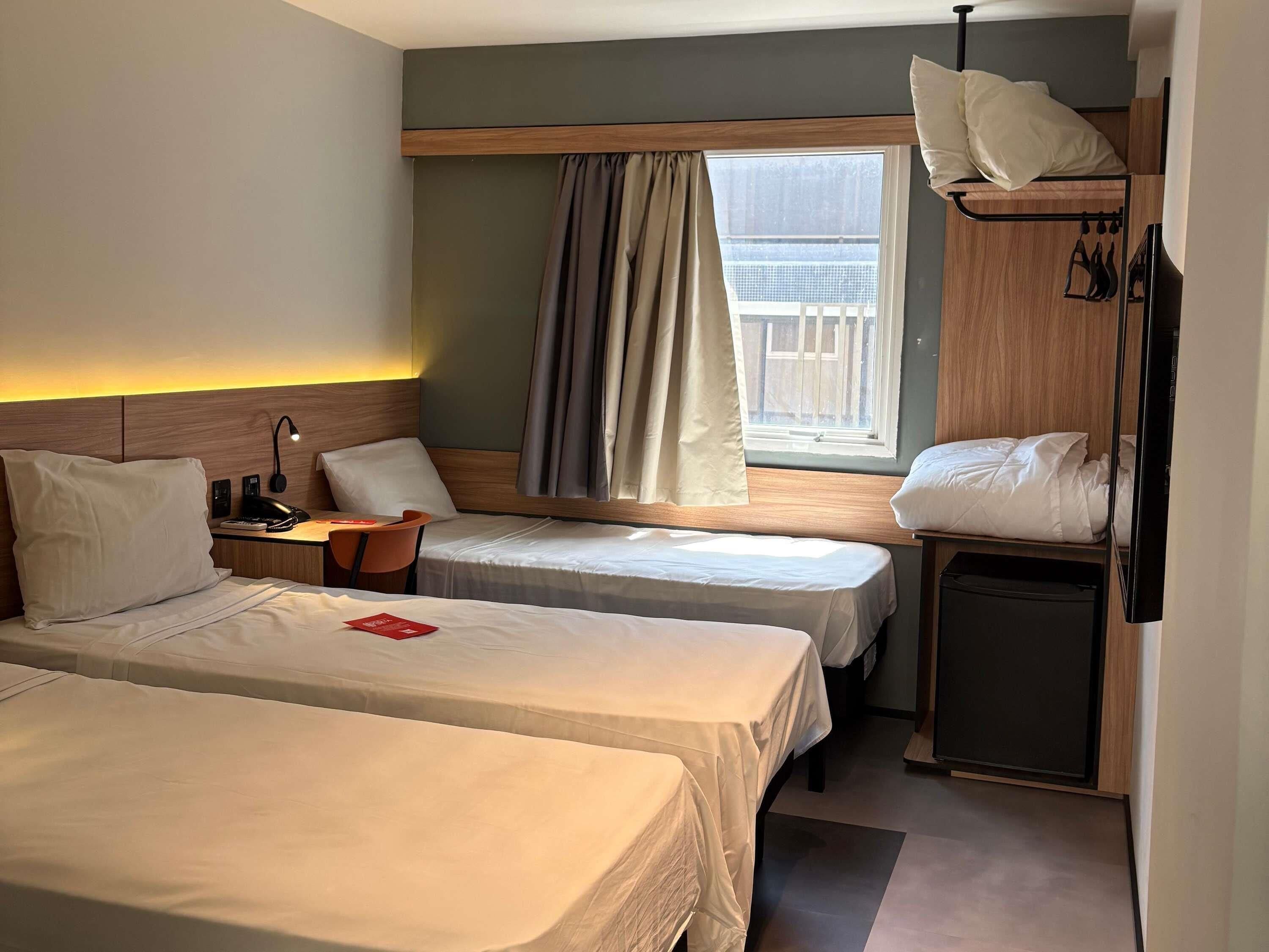 A hotel room with two beds, a desk, a chair, a TV, and a window with curtains.
