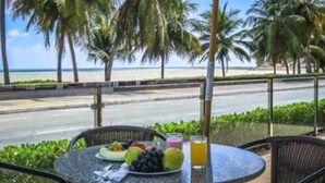A table with a fruit plate, a glass of juice, and a plate of food, overlooking a beach with palm trees and a road. - Ibis Joao Pessoa (João Pessoa)