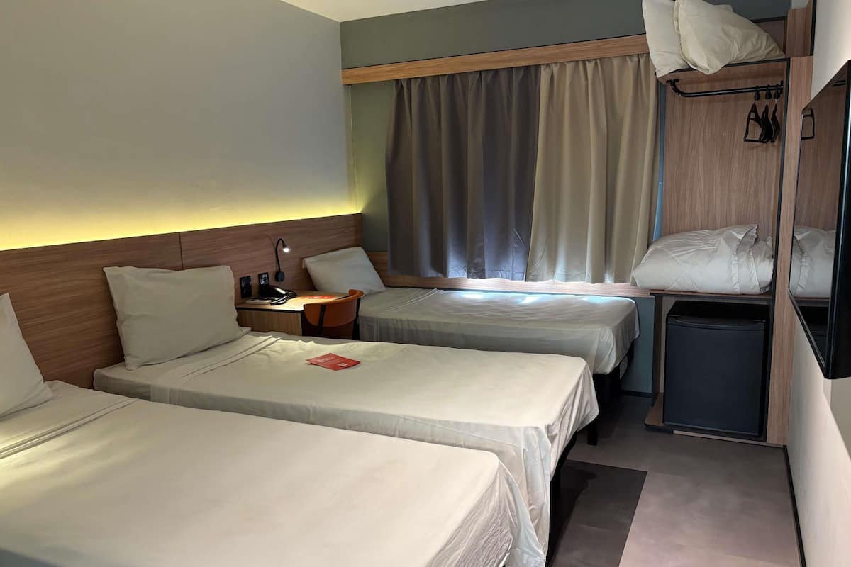 A hotel room with two single beds, a desk with a lamp, a small trash bin, and a window with curtains.