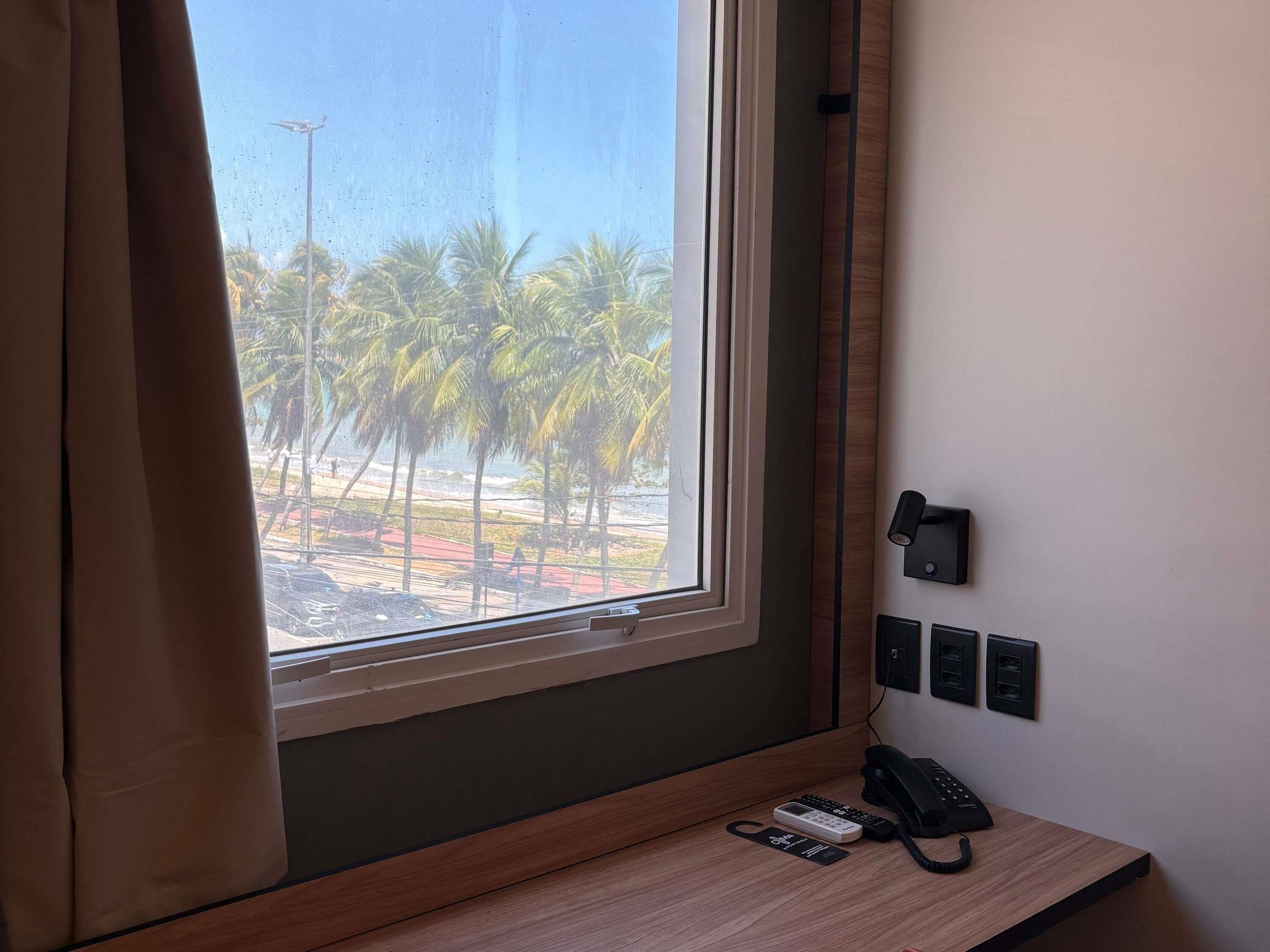 A hotel room with a wooden desk, a telephone, and a wall-mounted intercom. A window with curtains offers a view of palm trees and a beach.