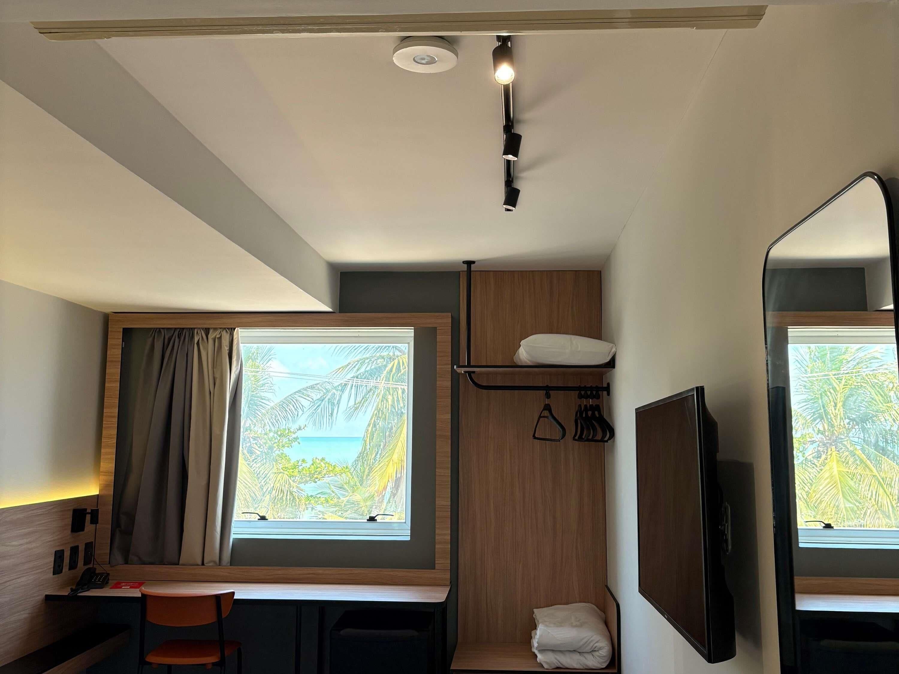 Ein kleines Hotelzimmer mit Schreibtisch, Stuhl, Bett, Wand-Fernseher, Fenster mit Vorhängen und einer Deckenleuchte.
