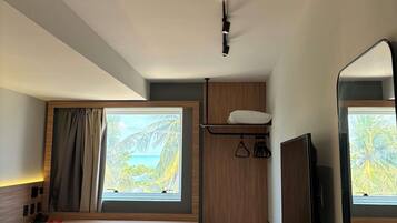 Ein kleines Hotelzimmer mit Schreibtisch, Stuhl, Bett, Wand-Fernseher, Fenster mit Vorhängen und einer Deckenleuchte.