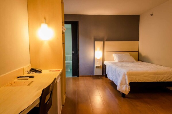 Habitación de hotel con cama, mesita de noche y escritorio con silla. Se ve una puerta que da al baño.